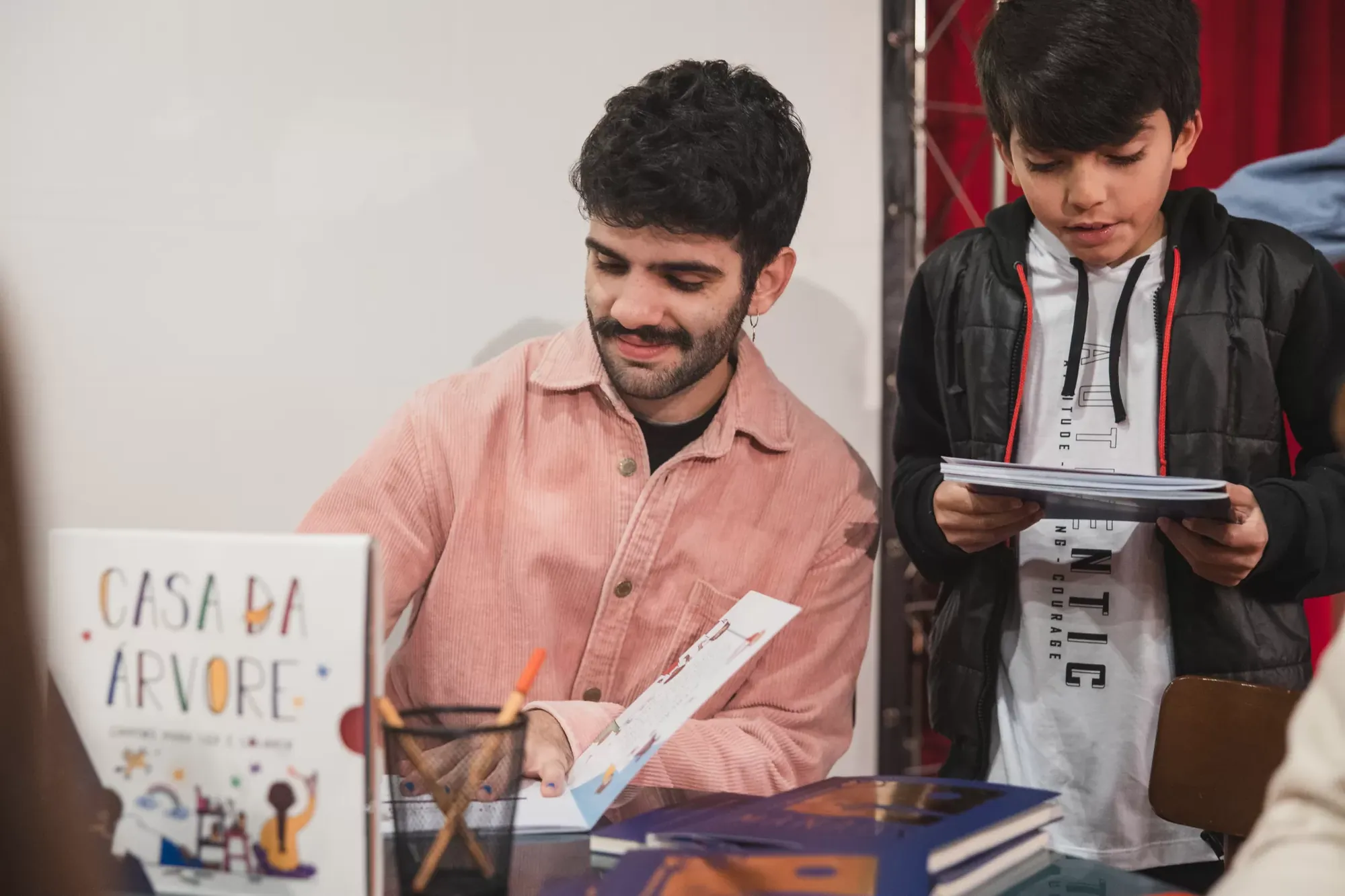 Menino lendo ao lado de um dos autores, em uma mesa de livros